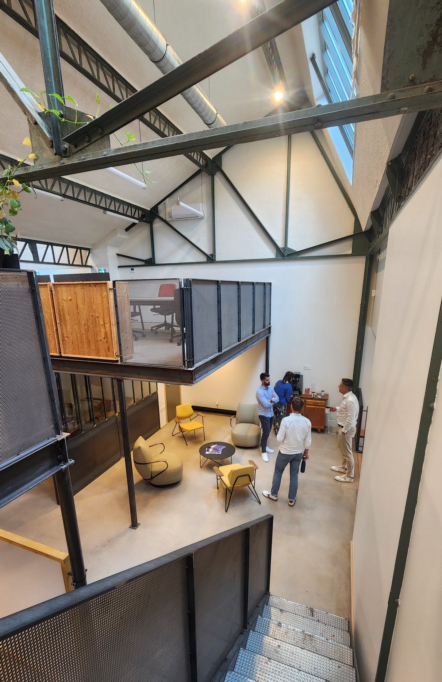 Vue depuis l'escalier de l'espace de coworking NOO à Oullins (Lyon Sud), montrant des coworkers qui échangent dans l'espace de vie au style industriel et la mezzanine, à 100m du Métro B.